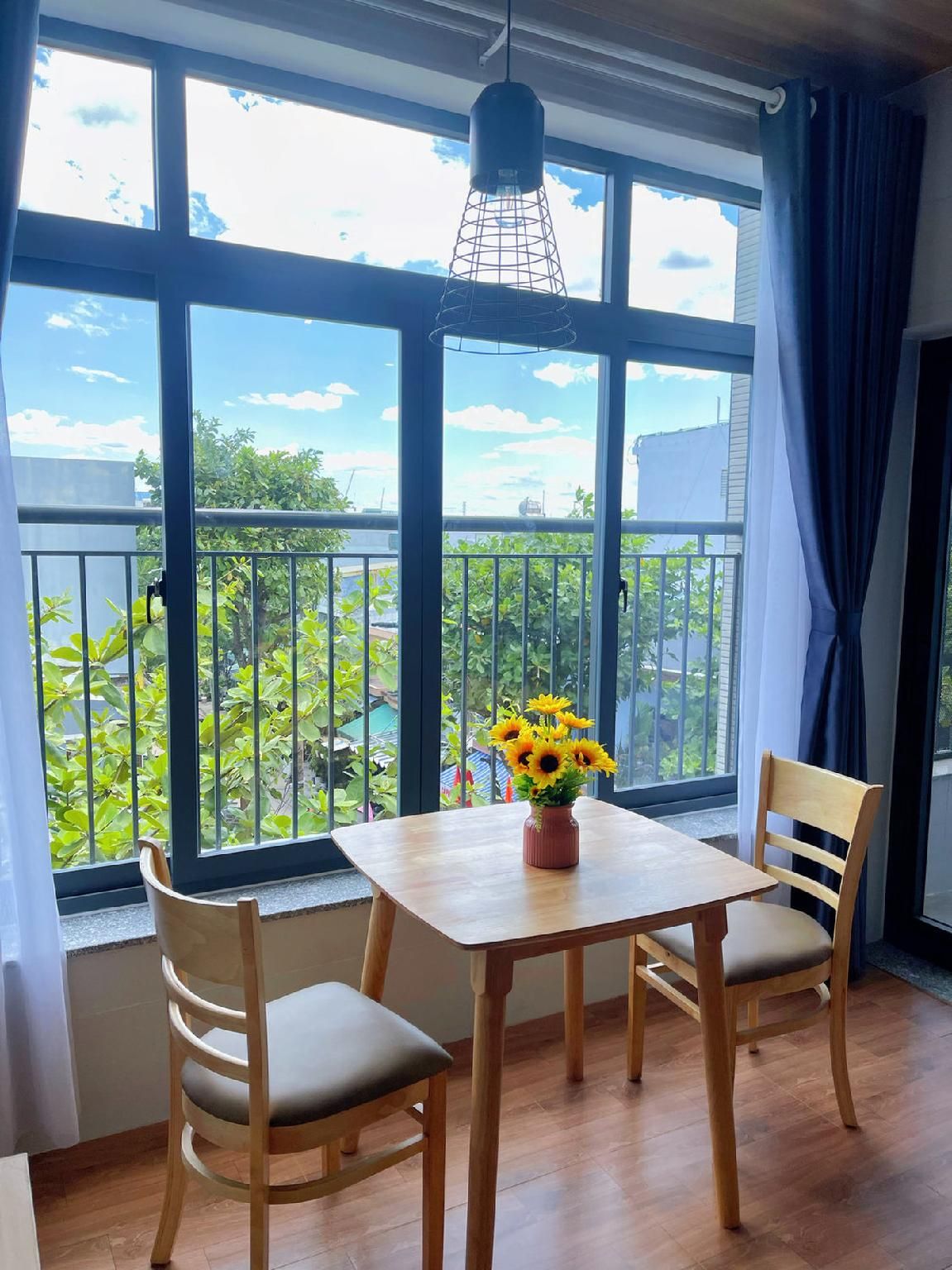 A room with a wooden table and chairs, a hanging light fixture, and large windows with a view of greenery.