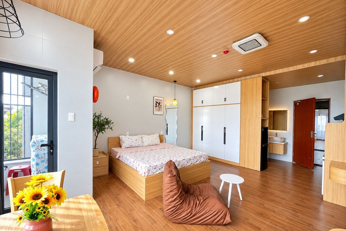 Une chambre moderne avec un plafond en bois, un lit recouvert d’une courtepointe à motifs floraux, une table de chevet, une commode, une plante en pot et une fenêtre donnant sur un beau paysage.