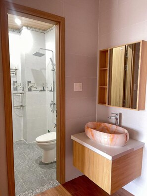 Une salle de bain comprenant des toilettes, un lavabo avec un receveur en marbre, une douche avec une porte en verre et un meuble en bois doté d’un miroir.