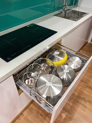 Una cocina con placa de inducción empotrada, un lavamanos de acero inoxidable y un escurreplatos con ollas y sartenes.