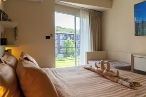 Une chambre d’hôtel avec un grand lit, un lit plus petit, un bureau et une vue sur un immeuble et la verdure environnante.