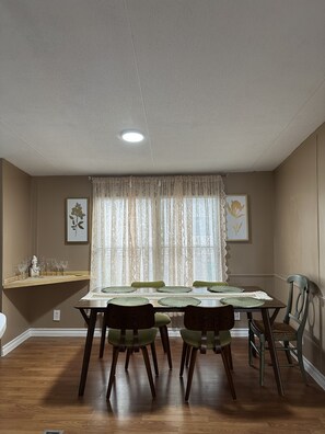 Uma sala de jantar com uma mesa posta para quatro pessoas, ladeada por duas cadeiras, uma janela com cortinas de renda e duas fotos emolduradas nas paredes.