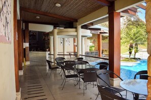 Un espace extérieur aménagé en terrasse, avec des tables et des chaises rondes, une piscine en arrière-plan et une allée couverte.