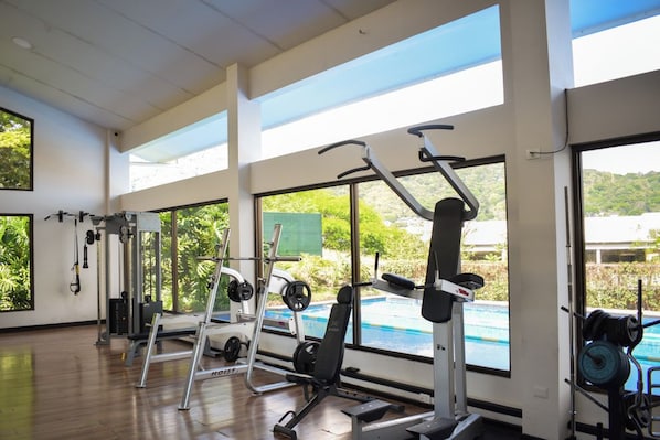 Une salle de sport équipée de divers appareils d’exercice, dotée de grandes fenêtres et offrant une vue sur une piscine.