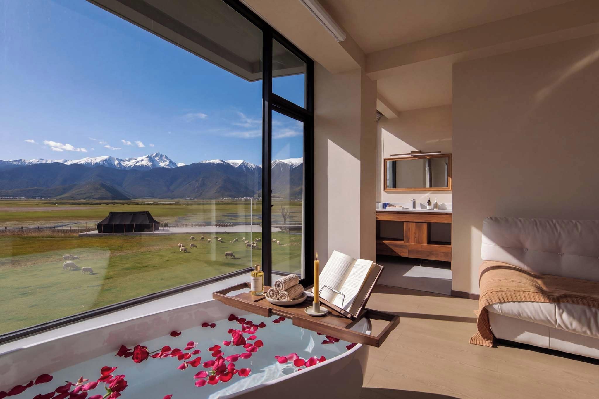 Ein modernes Badezimmer mit einer großen Badewanne, gefüllt mit Wasser und Rosenblüten, einer Holztablett mit Pflegeprodukten und Blick auf eine Berglandschaft.