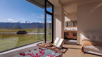 Ein modernes Badezimmer mit einer großen Badewanne, gefüllt mit Wasser und Rosenblüten, einer Holztablett mit Pflegeprodukten und Blick auf eine Berglandschaft.