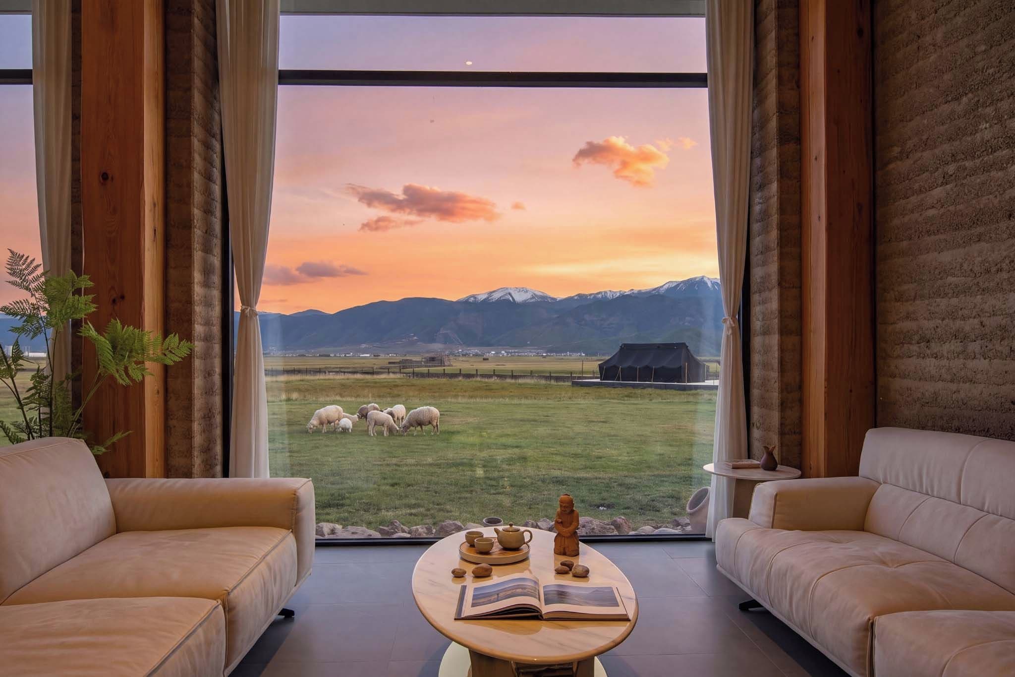 A room with a large window overlooking a grassy field with sheep, a mountain range, and a building in the distance. There are two sofas and a small round table with a book and a tea set.