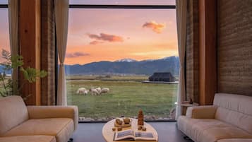 A room with a large window overlooking a grassy field with sheep, a mountain range, and a building in the distance. There are two sofas and a small round table with a book and a tea set.