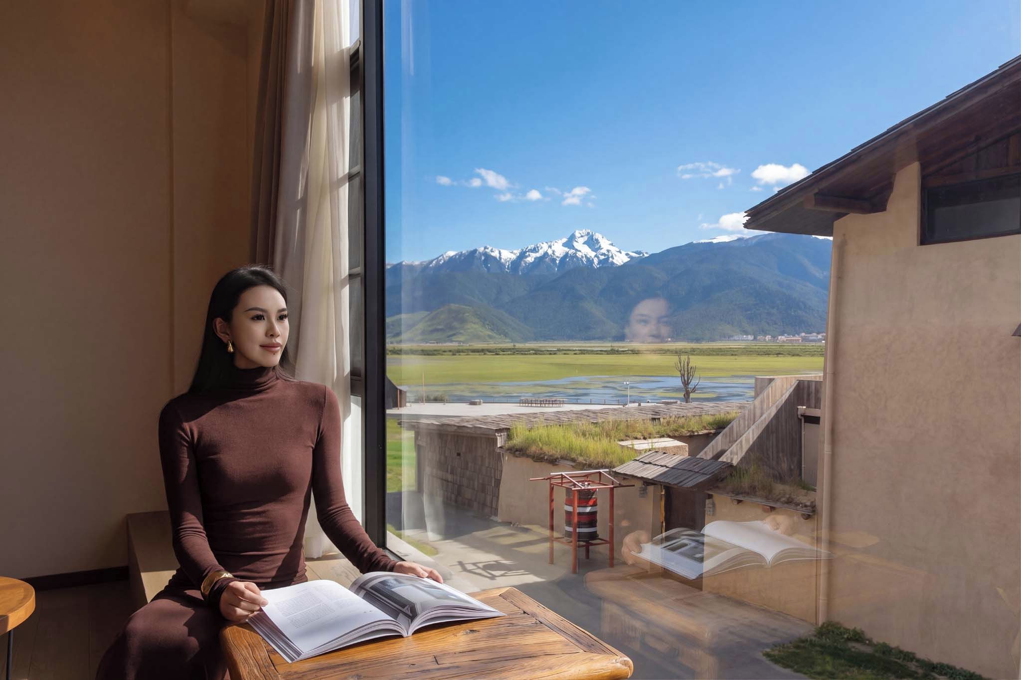 Una persona sentada a una mesa con un libro abierto, contemplando un paisaje montañoso a través de un amplio ventanal.