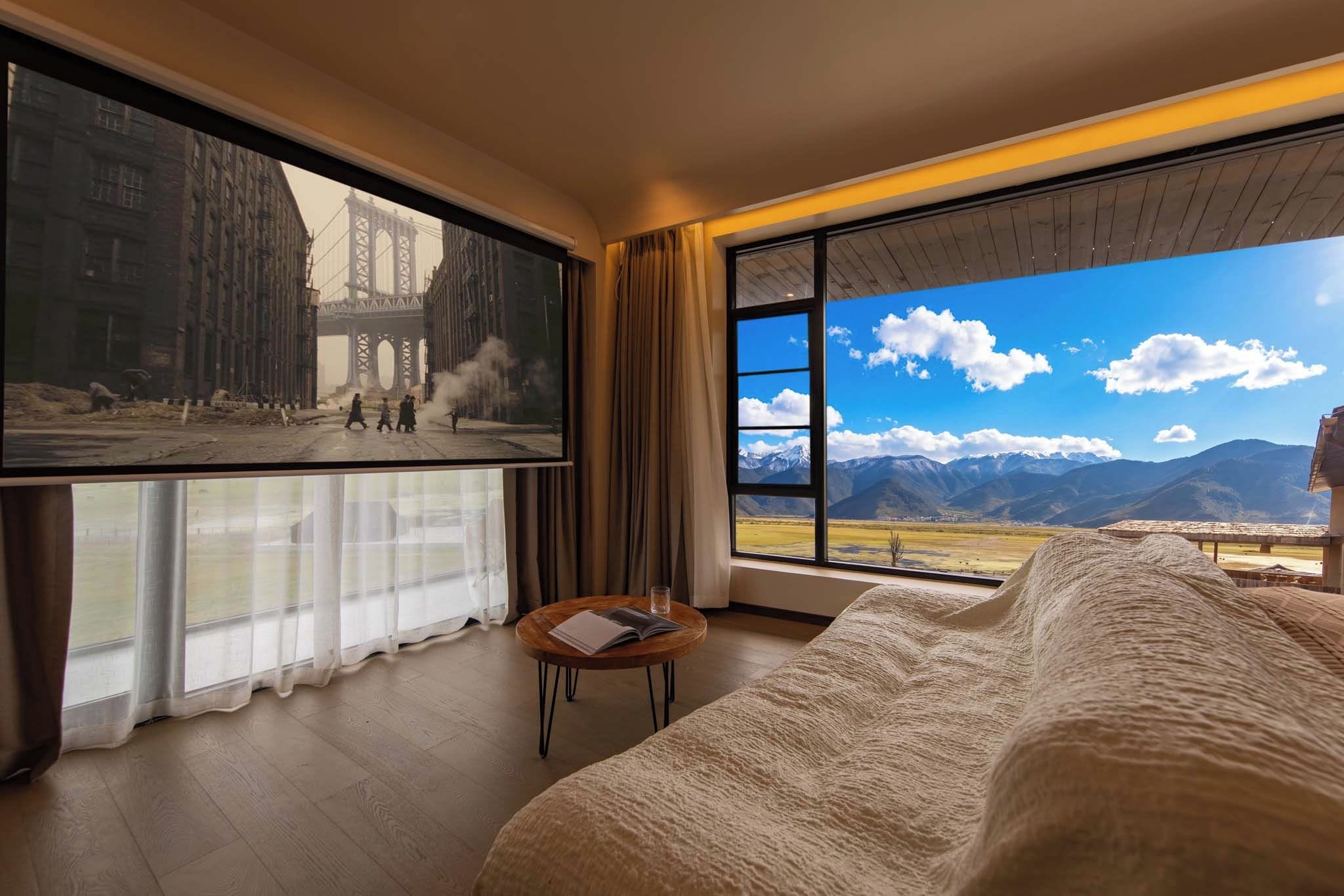 Ein Hotelzimmer mit einem großen Bett, einem kleinen Tisch mit einem Buch, einem Fernsehbildschirm, der eine Stadtansicht zeigt, und einem großen Fenster mit Blick auf die Berge.