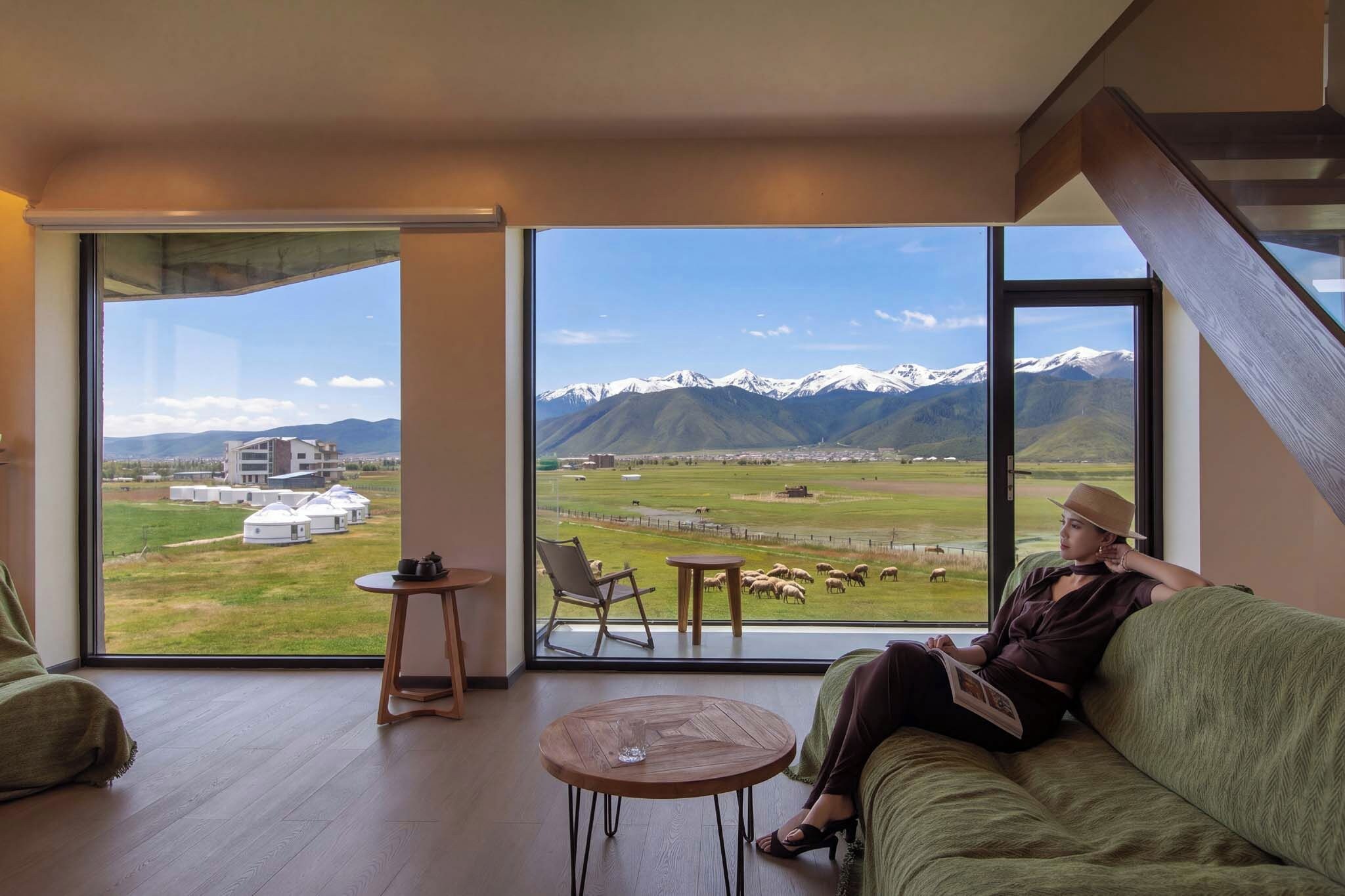 A room with a large window offering a mountain view, a green sofa, a small table, and a chair.