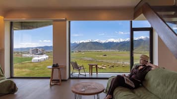 A room with a large window offering a mountain view, a green sofa, a small table, and a chair.