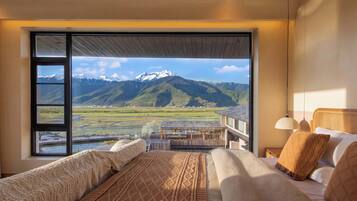 A bedroom with a large window offering a mountain view, a bed with patterned bedding, and a wooden ceiling.