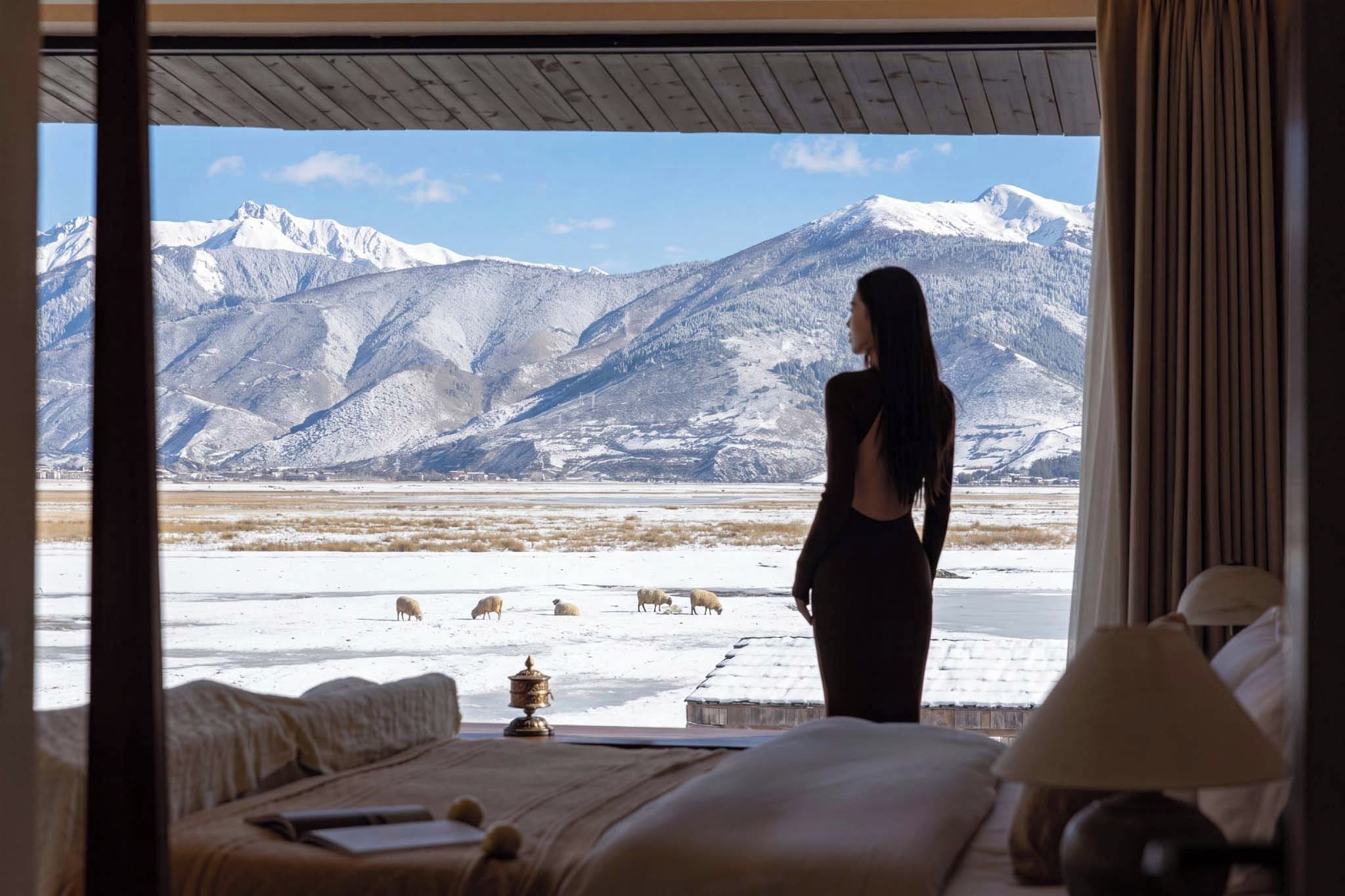 Una habitación con cama, lámpara y vistas a un paisaje nevado con montañas y ovejas pastando.