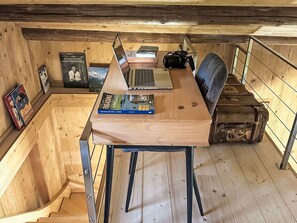 Ein Holztisch mit einem Laptop, Büchern und einer Kamera in einem raumähnlichen Blockhaus mit Holzwänden und einer Treppe.
