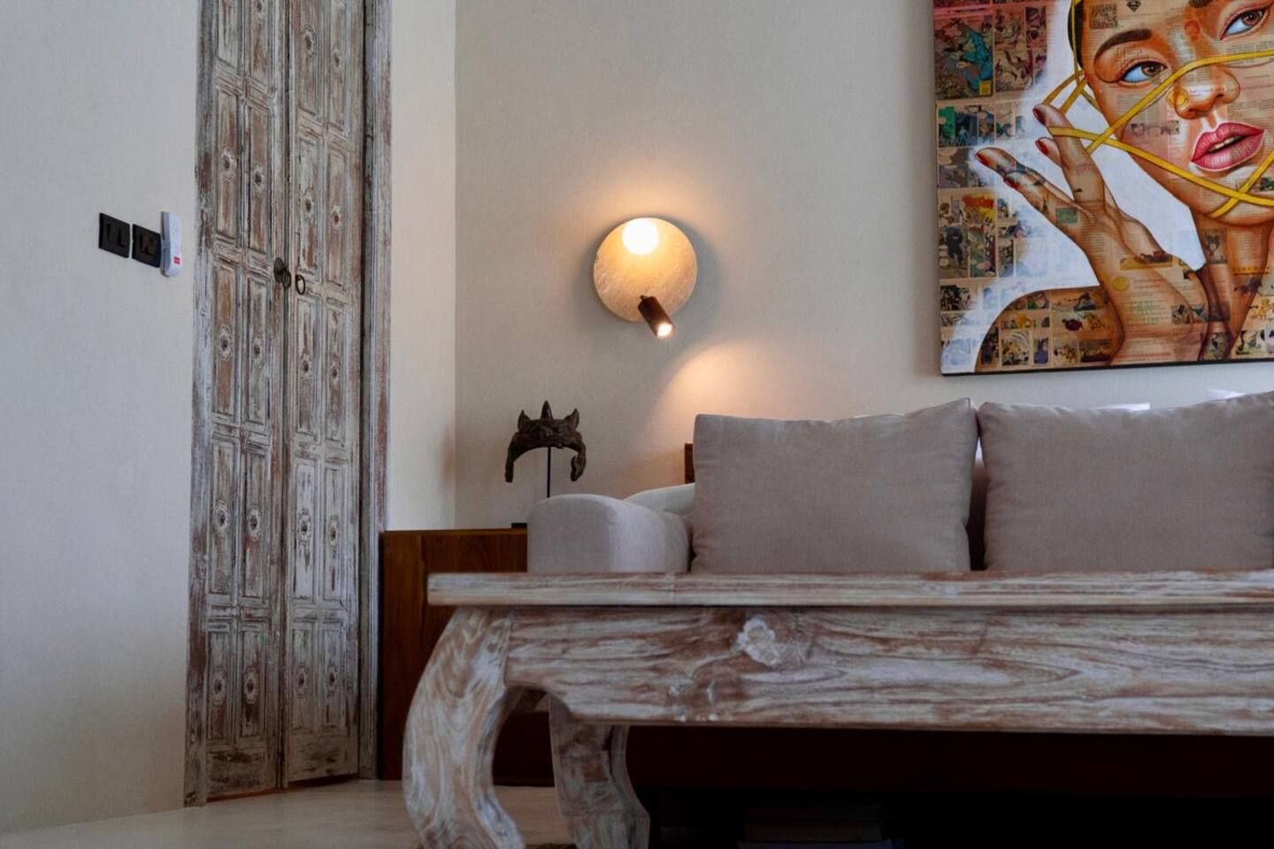 A room with a bed, a wall-mounted light, a painting, and a rustic wooden table.