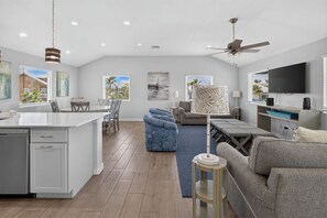 A modern kitchen with a central island, a dining area with chairs, a living room with a sofa and a coffee table, and a television mounted on the wall. - Fluorescent Beauty 4BR Home (Port Aransas)
