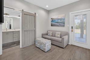 Um interior moderno com um sofá cinzento, uma porta de correr, um quadro de um cais e um banquinho pequeno com tema oceânico.