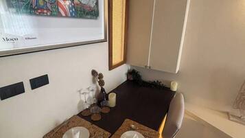Un coin repas avec une table dressée pour deux personnes, une œuvre d’art encadrée au mur et une fenêtre avec des stores.