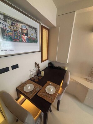 Een eethoek met een gedekte tafel voor twee personen, een ingelijst kunstwerk erboven en een raam met luxaflex.