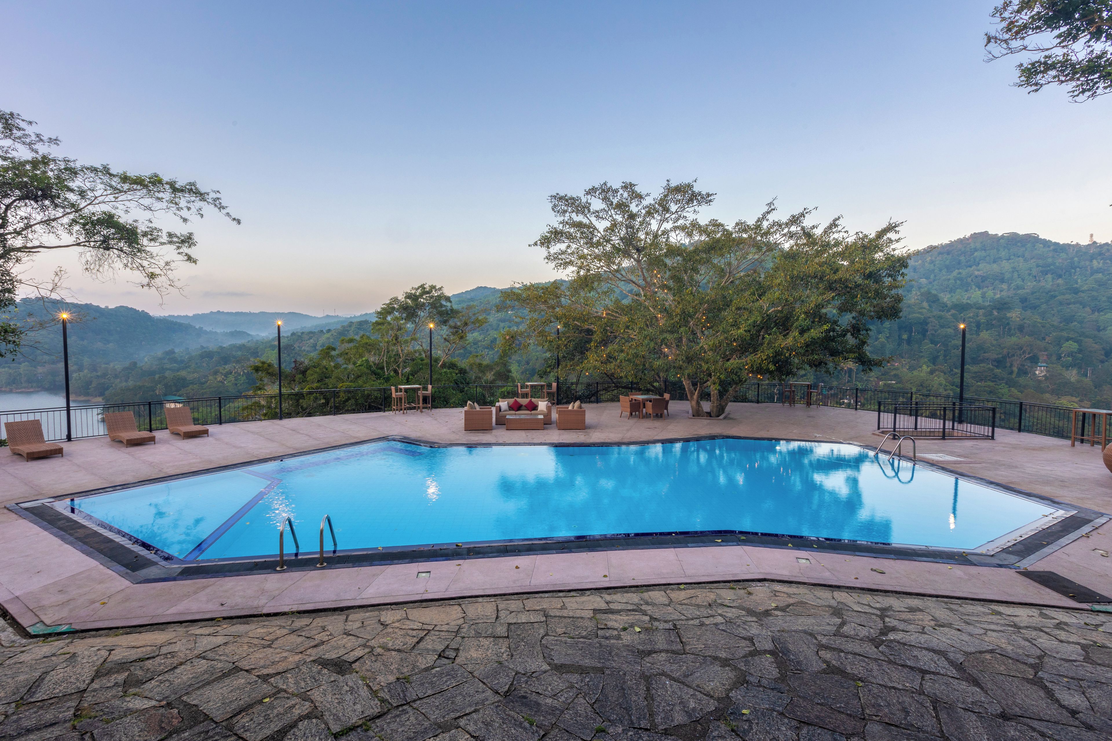 A swimming pool with a stone border, surrounded by lounge chairs and tables, set against a backdrop of hills and trees.