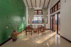 A room with a green wall, a large window with curtains, a wooden table with chairs, and a potted plant. - Indra Manel Family Holiday Resort (Kandy)