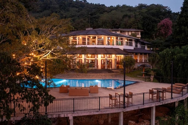 A large, multi-story building with a swimming pool, outdoor seating, and a well-lit balcony area.