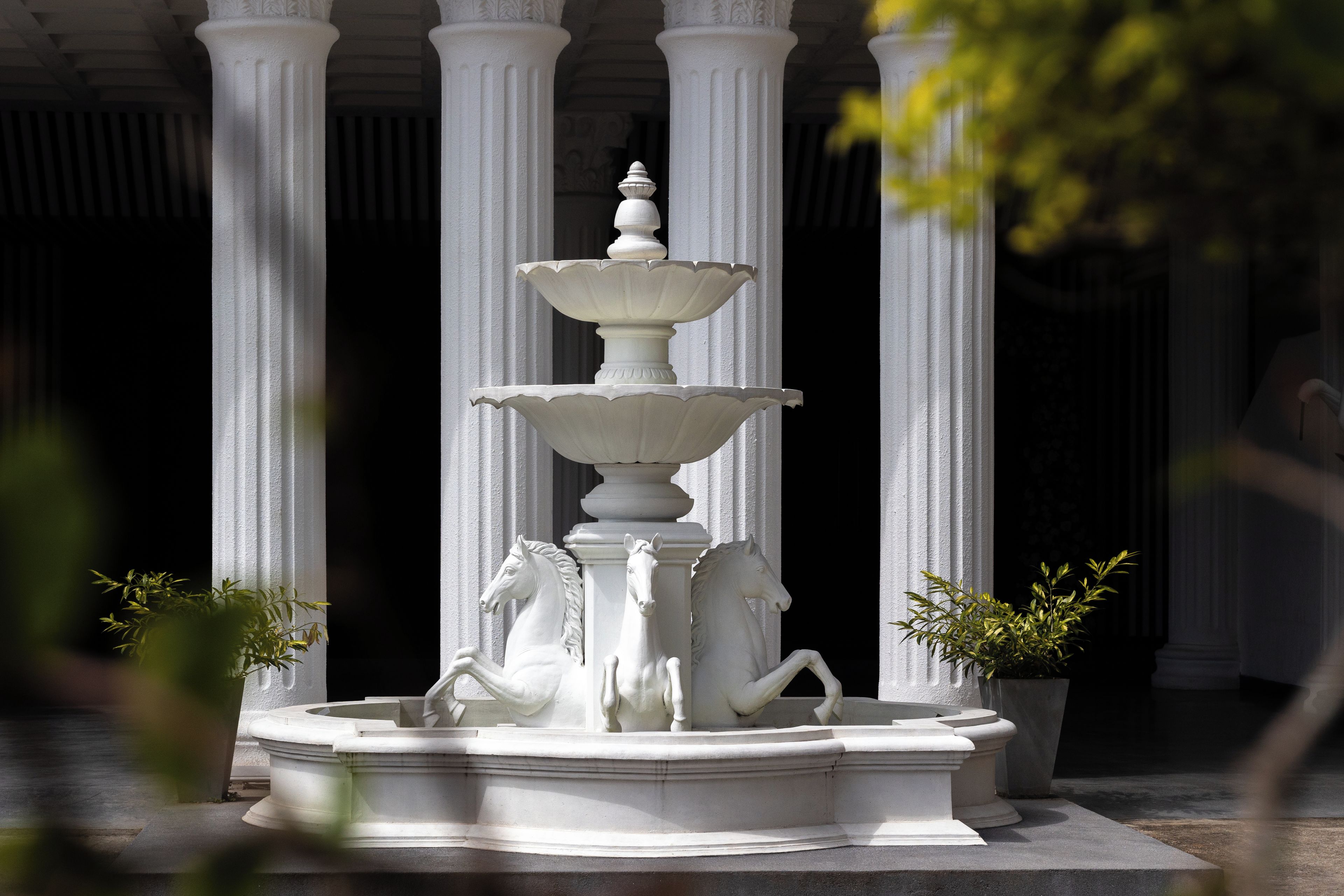 A white stone fountain with multiple tiers and horse statues, surrounded by white columns.