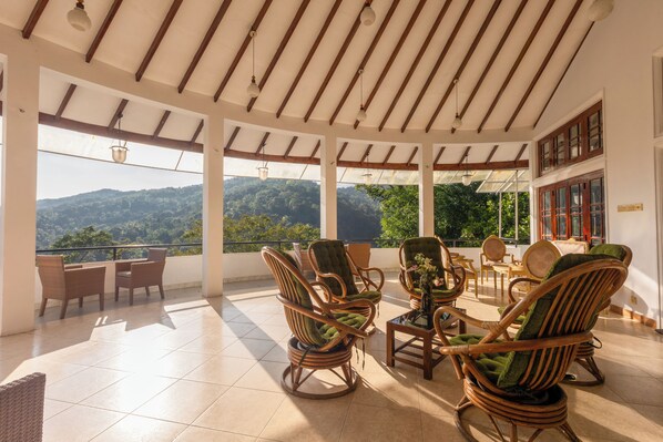 A covered outdoor seating area with wicker furniture, large windows, and a view of a mountainous landscape.
