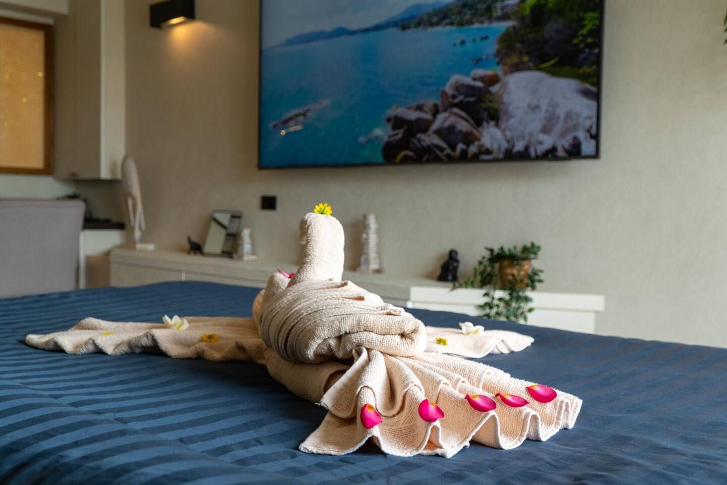Un lit bien fait, avec une serviette disposée en forme de cygne, un tableau représentant un paysage côtier accroché au mur et une petite statue posée sur la table de chevet.