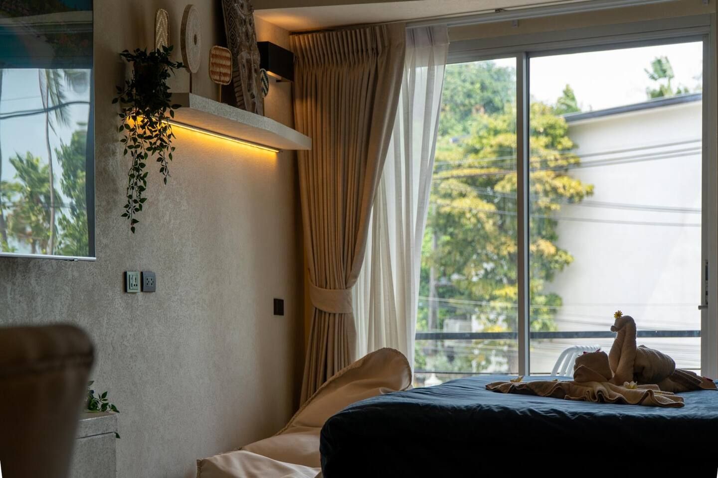 Une chambre à coucher avec un lit, une fenêtre avec des rideaux, un luminaire fixé au mur et une plante.