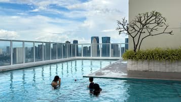 Una piscina en la azotea con vistas a un horizonte urbano, rodeada de barandillas de vidrio y un pequeño árbol.
