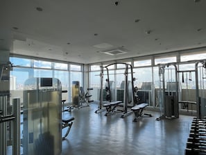 Une salle de sport équipée de divers appareils d’exercice, dotée de grandes fenêtres et offrant une vue sur la ville.