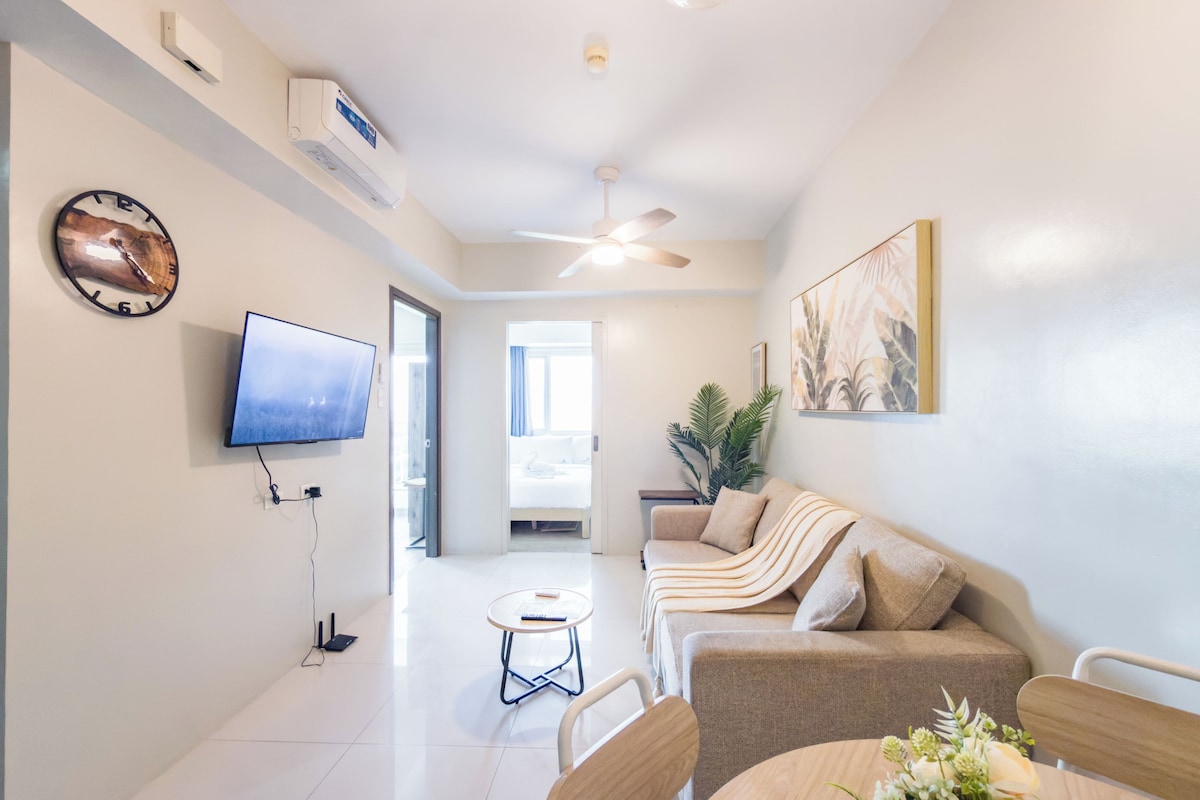 Una sala de estar moderna con un televisor de pantalla plana, un ventilador de techo, un sofá, una mesa de comedor con sillas y un reloj montado en la pared.