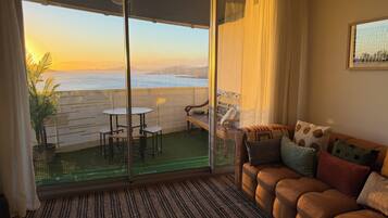 Ein Zimmer mit einer Schiebetür zu einem Balkon mit Tisch und Stühlen, einer Couch mit Kissen und Blick auf das Meer.