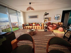 A modern living room with a large sofa, a coffee table, and striped chairs. There is a large window with a view of a balcony. - Sarcelle - Noumea 18th Floor Refined Apartment with Sea and Mountain views (Nouméa)