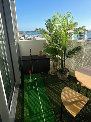 A small balcony with a putting green, a table, and a potted palm tree. - Sarcelle - Noumea 18th Floor Refined Apartment with Sea and Mountain views (Nouméa)