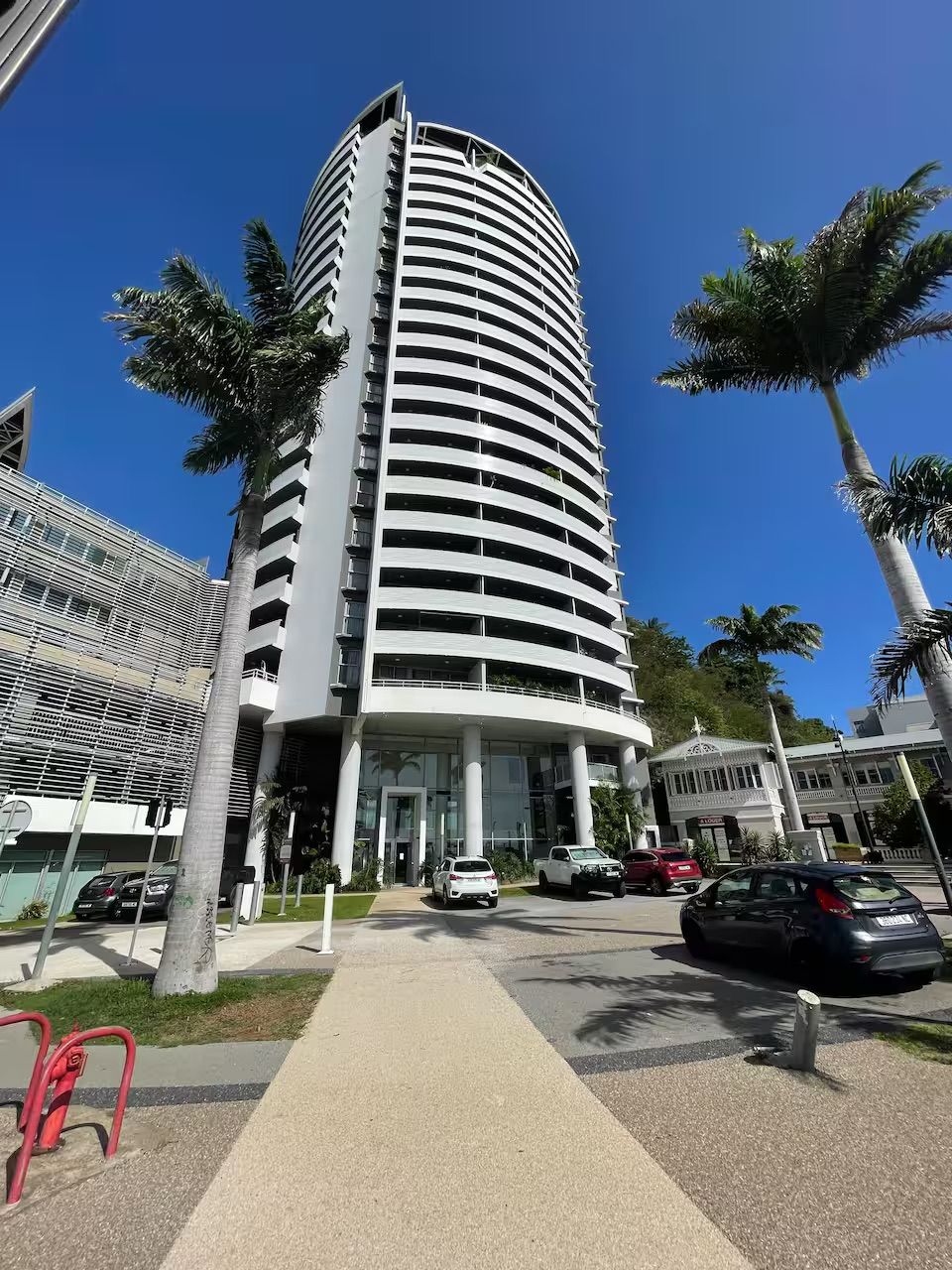 A tall, cylindrical building with multiple balconies, surrounded by palm trees and parked cars.