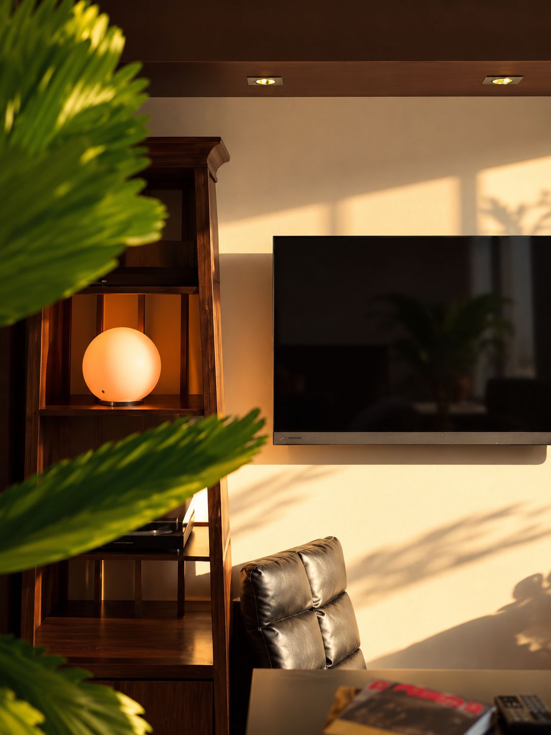 A modern living room with a flat-screen TV mounted on the wall, a leather sofa, a wooden entertainment unit with a spherical lamp, and a coffee table with a remote control.