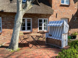 Ein Haus mit Backsteinen und einem Strohdach, ein Holztisch, zwei Stühle und ein gestreifter Sonnenschirm.
