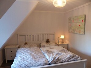 Ein Schlafzimmer mit schrägem Dach, einem Bett mit gemusterten Betttextilien, einer Nachttischlampe, einem Nachttisch und einem Blumenbild an der Wand.