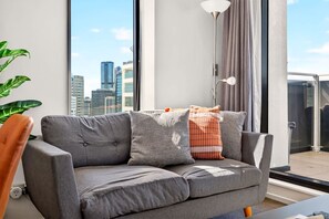 A modern living room with a grey sofa, a large window with a city view, and a floor lamp. - Cosy 2BR in the heart of CBD + Balcony Zodiak Stay (Auckland)