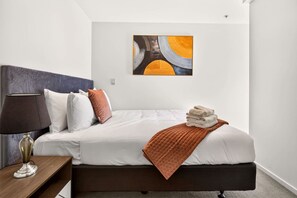 Una habitació d'hotel amb un llit, una cambrera de nit, una làmpada, una obra d'art a la paret i una tovallola doblegada sobre el llit.