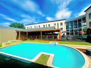 A swimming pool with a wooden deck and a covered seating area, surrounded by a well-maintained lawn and a building with large windows.