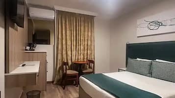 Quarto de hotel com cama, escrivaninha, cadeira, mesa pequena e televisão fixada na parede.