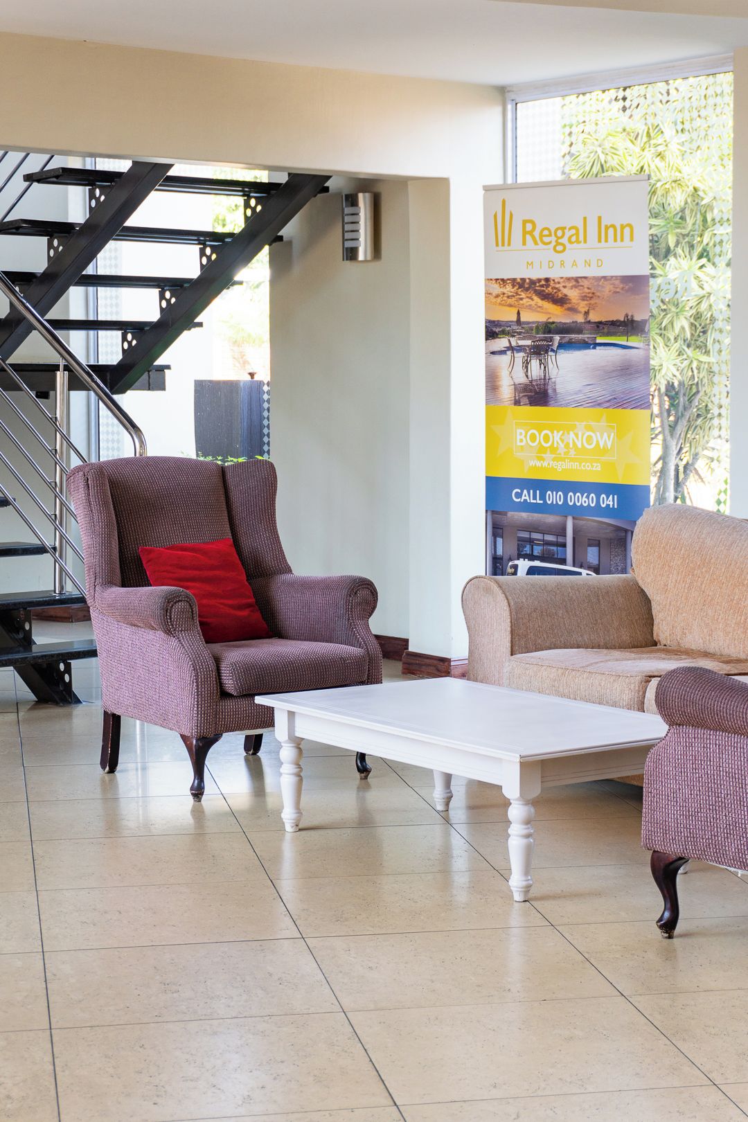 Un hall de hotel con una escalera, dos sillones, una mesa de centro blanca y un cartel promocional para Regal Inn.