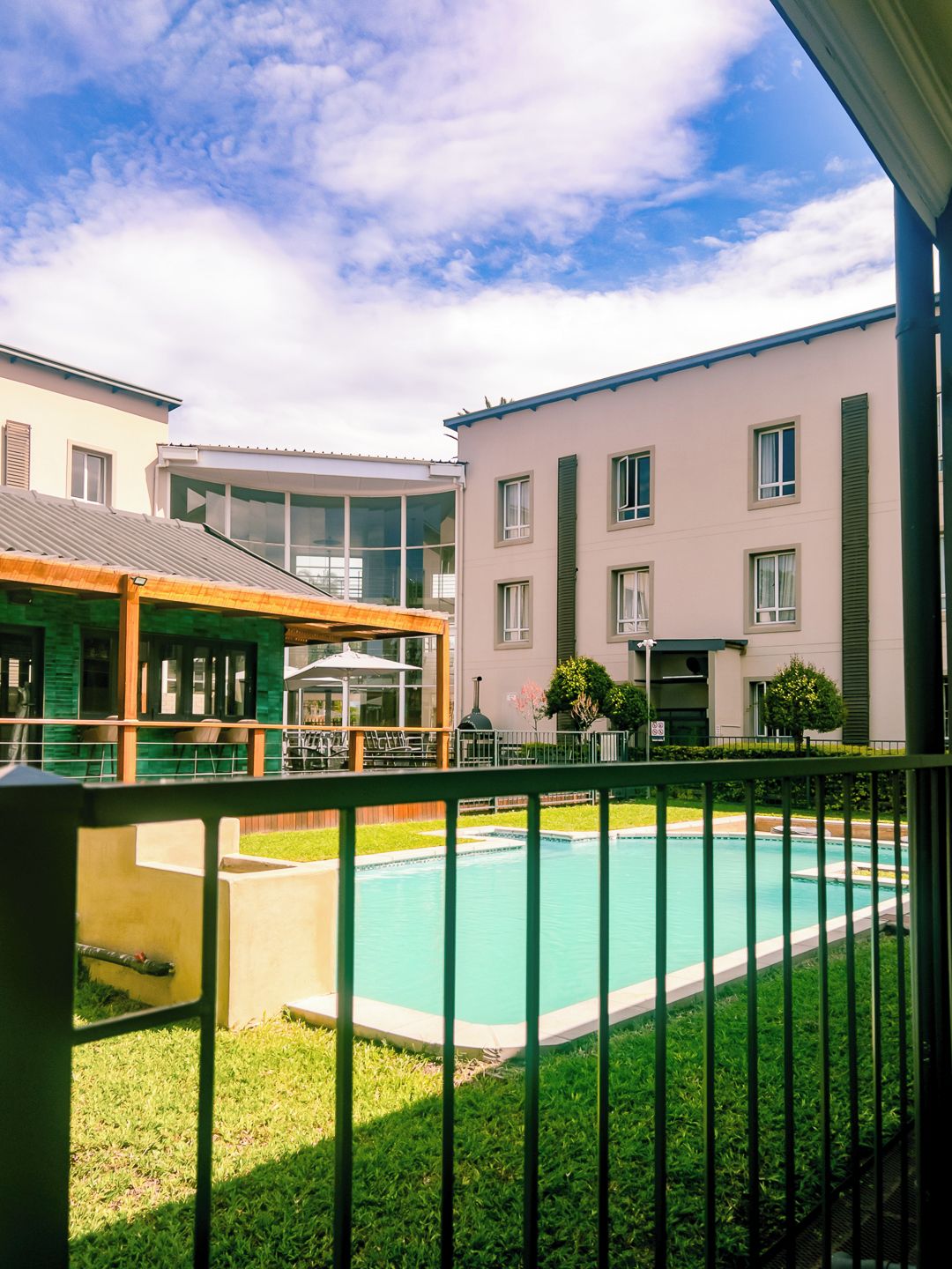 Una piscina con valla verde, un área de estar techada al aire libre y edificios al fondo.