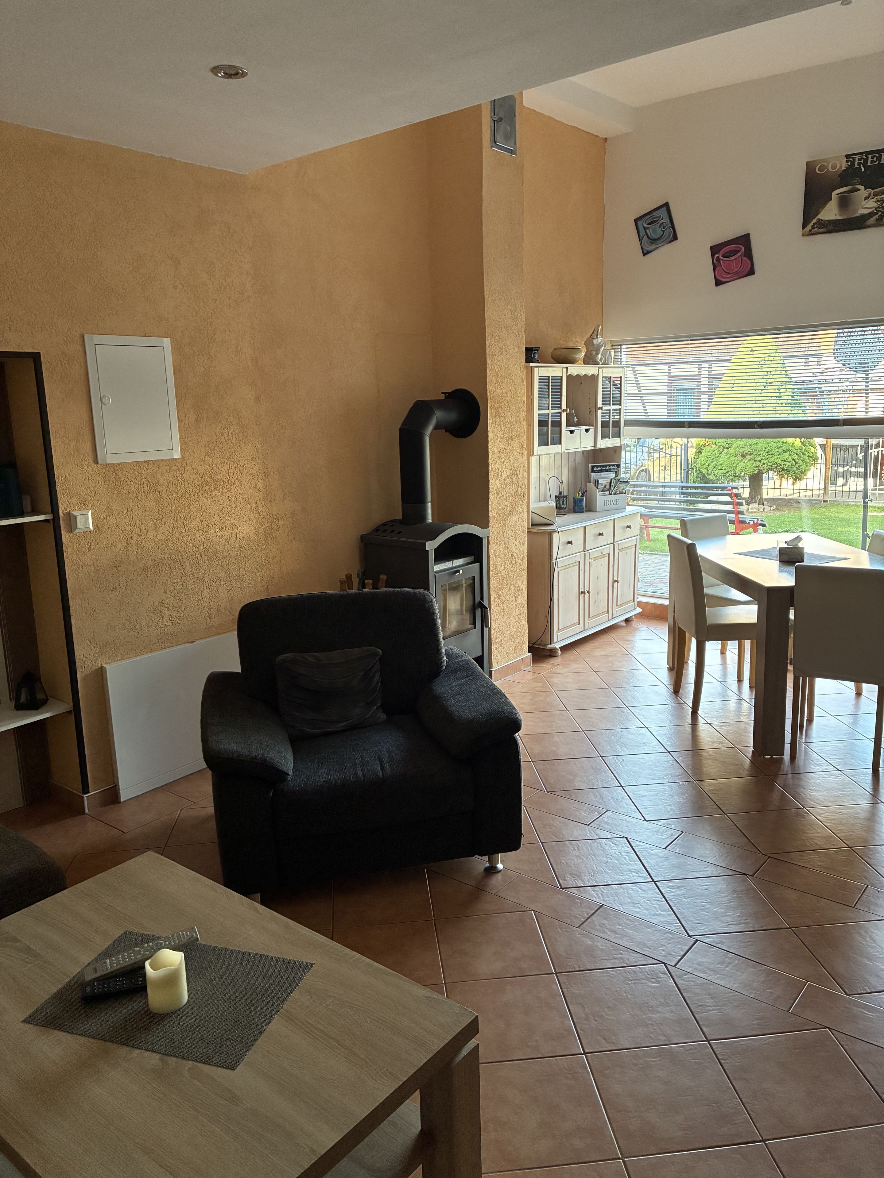 A living room with a sofa, a table with a remote control and a candle, a kitchen area with a stove, and a dining area with chairs.