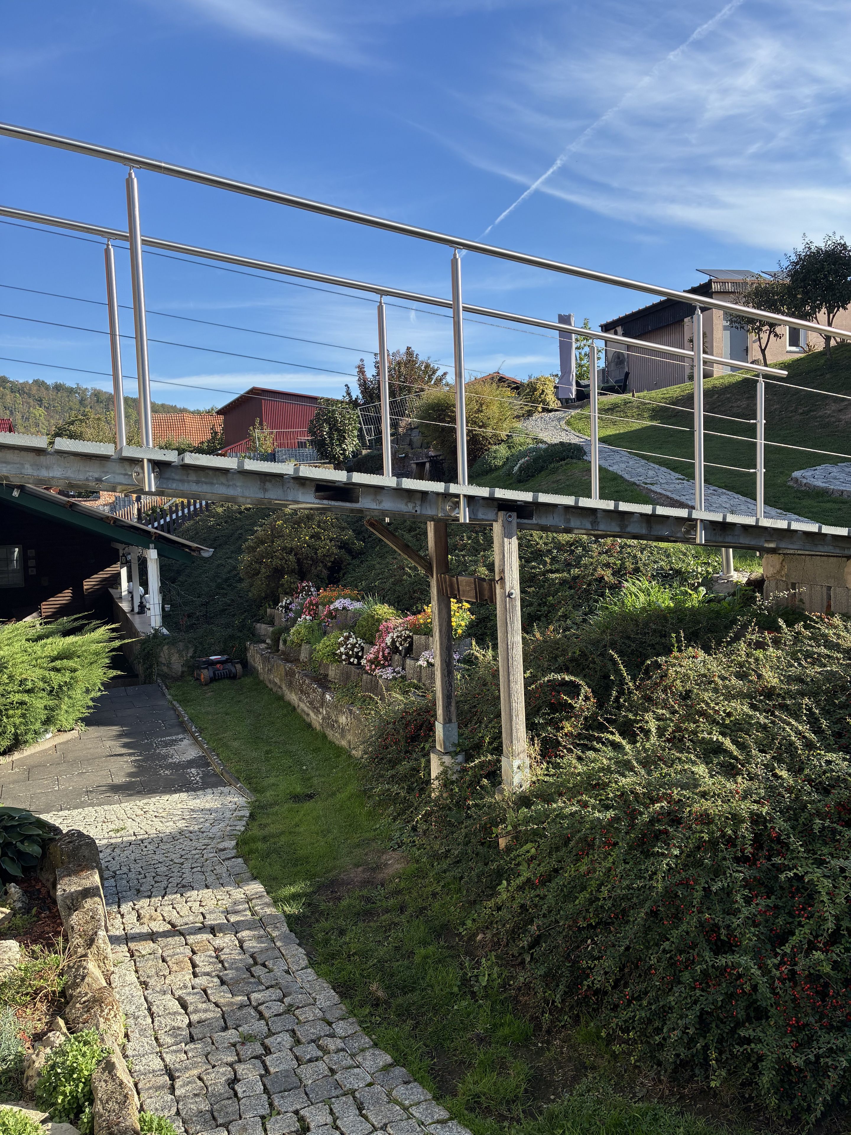 Un pont piétonnier avec des garde-corps en métal, un sentier pavé de pierres et une colline verdoyante, avec un bâtiment visible au loin.