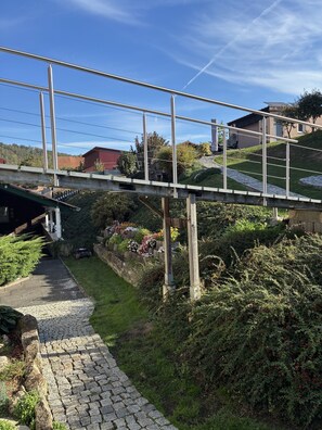 Eine Fußgängerbrücke mit Metallgeländern, einem Steinweg und einem grasbewachsenen Hang mit einem Gebäude im Hintergrund.